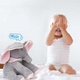 Une petite fille habillée en layette blanche à genoux en train de se caché les yeux, sur la gauche, une peluche éléphant gris et rose.