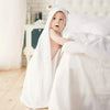 Un bébé debout au bord d'un lit, il porte un serviette cape blanche  sur la tête.